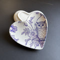 A heart-shaped ceramic dish with a white glaze and a blue-purple floral and owl pattern, shown on a dark gray background.