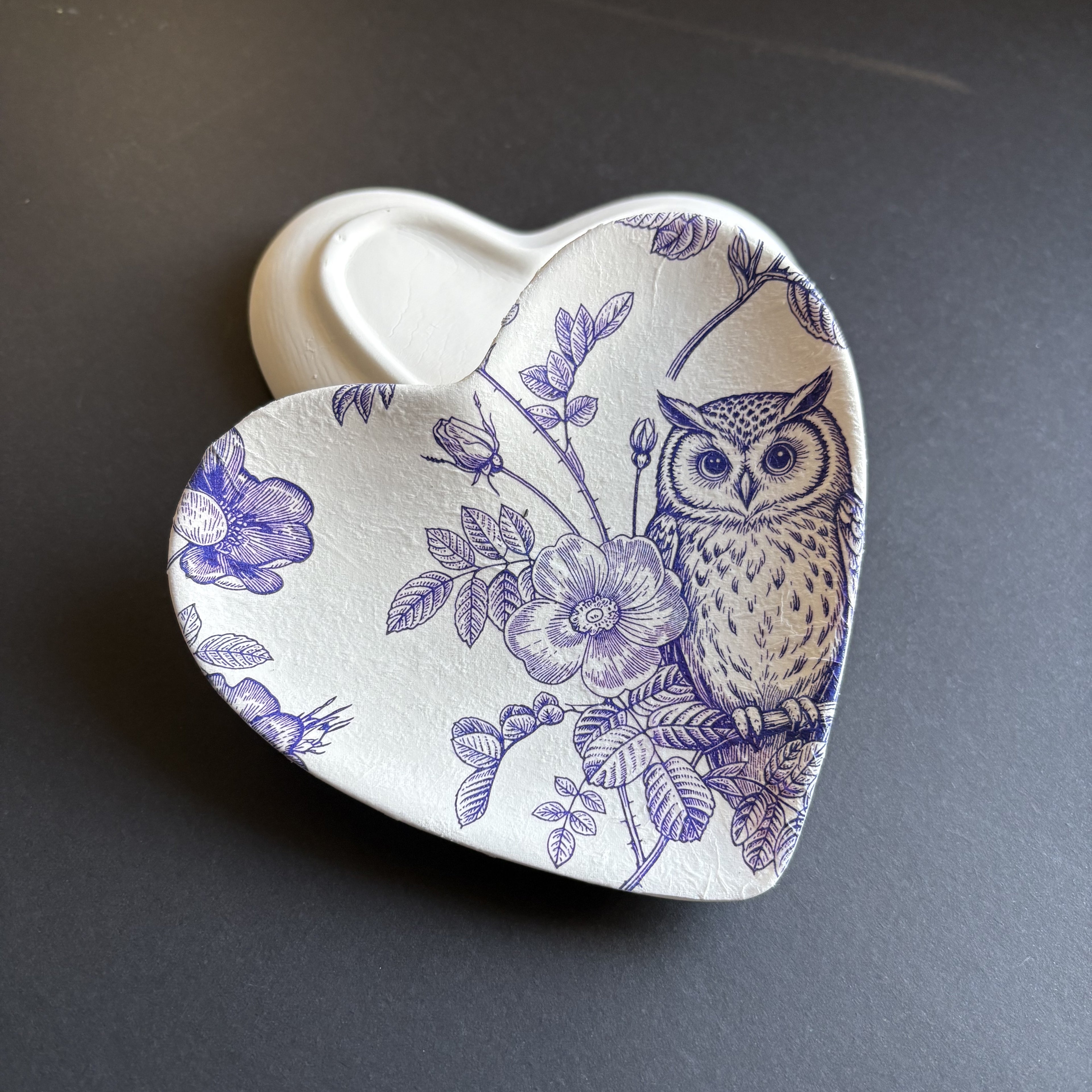 A heart-shaped ceramic dish with a white glaze and a blue-purple floral and owl pattern, shown on a dark gray background.