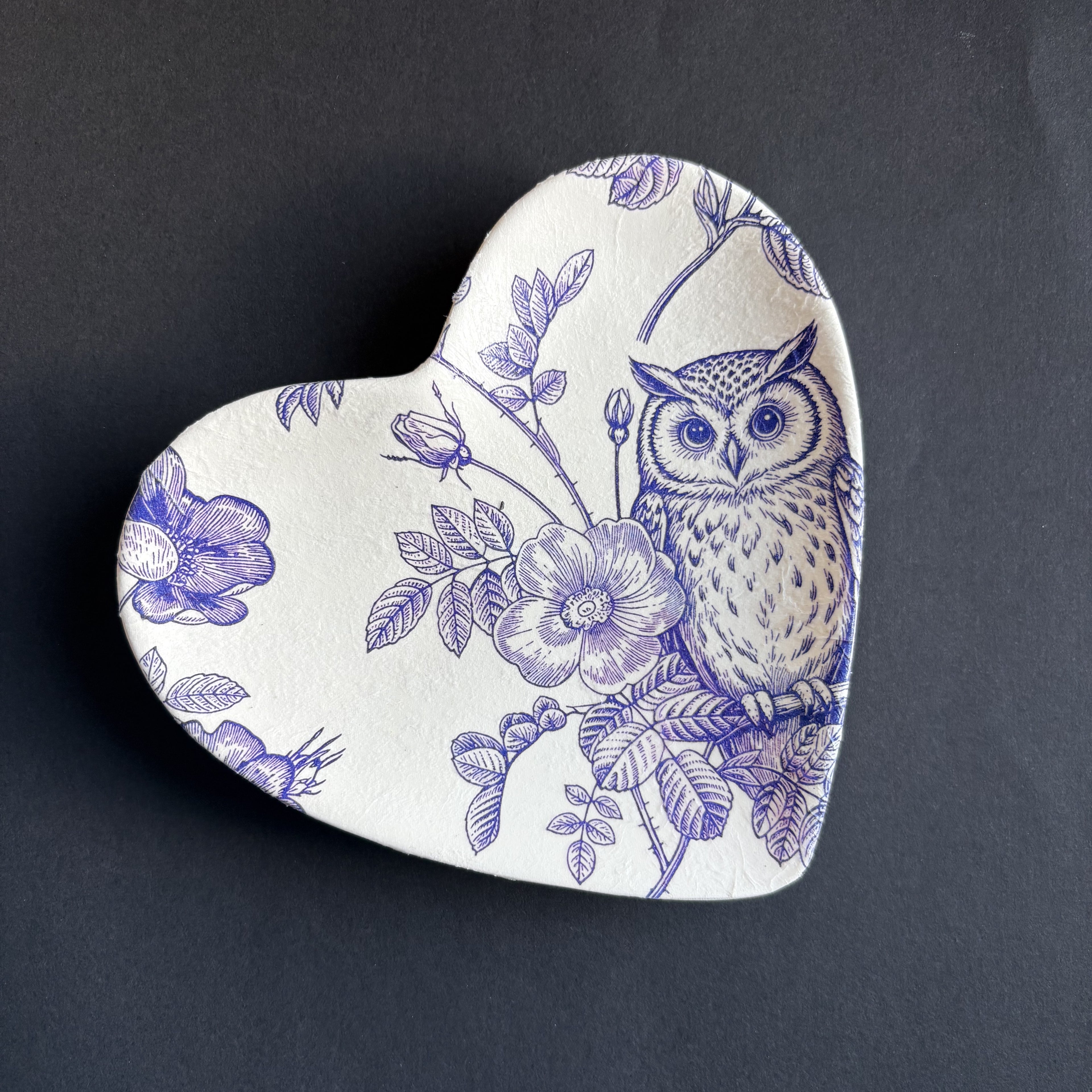 A heart-shaped ceramic dish with a blue floral and owl pattern, shown on top of a matching plain heart-shaped dish against a dark background.