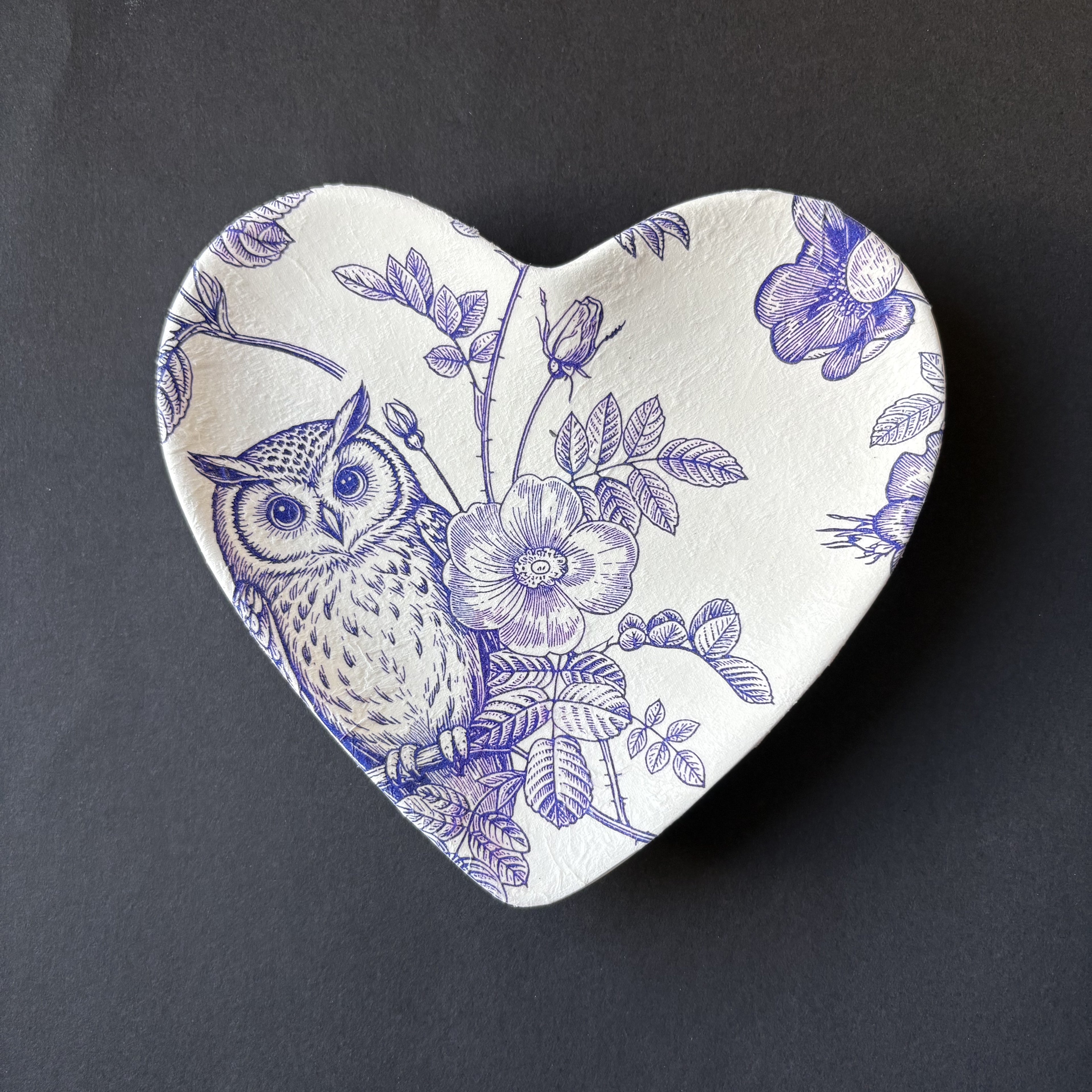 A heart-shaped ceramic dish featuring a blue and white floral and owl pattern, photographed on a dark gray background.