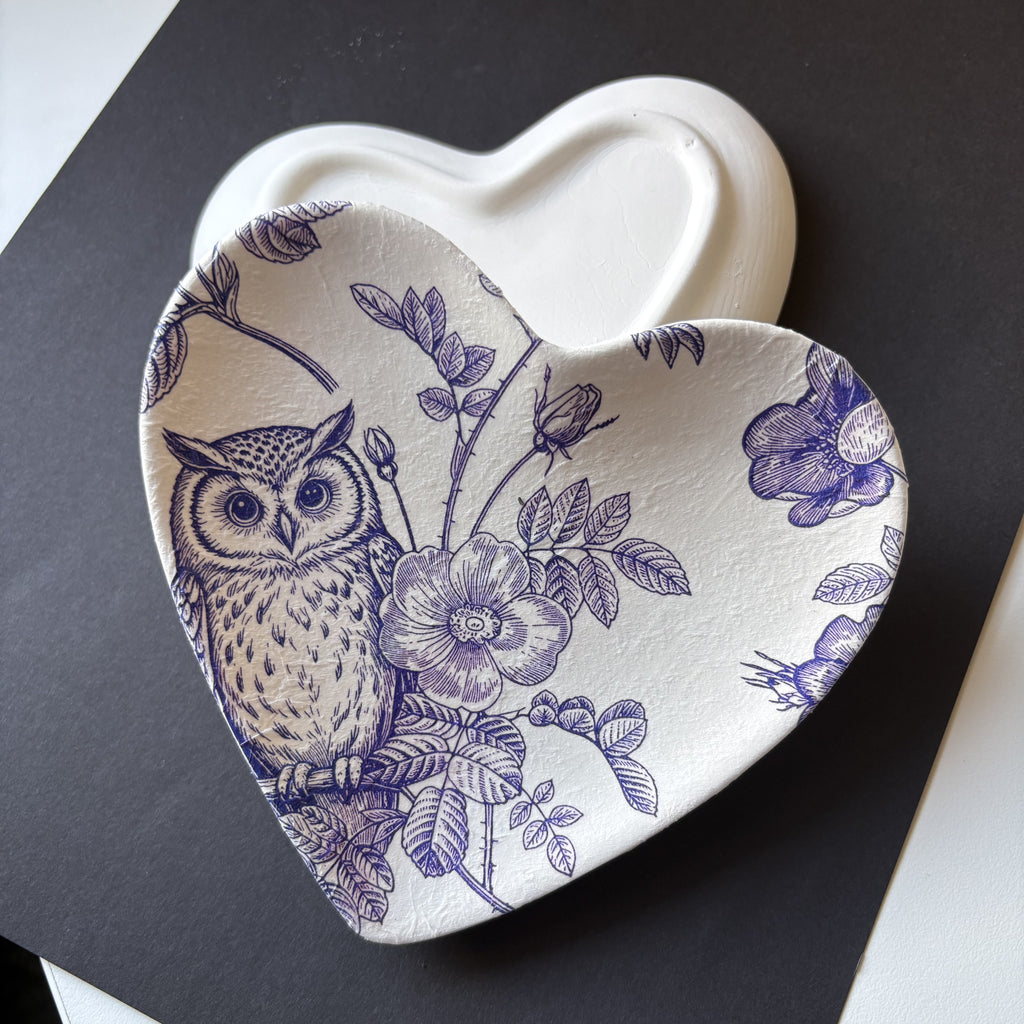 A heart-shaped ceramic dish with a blue floral and owl pattern, shown on top of a matching plain heart-shaped dish against a dark background.