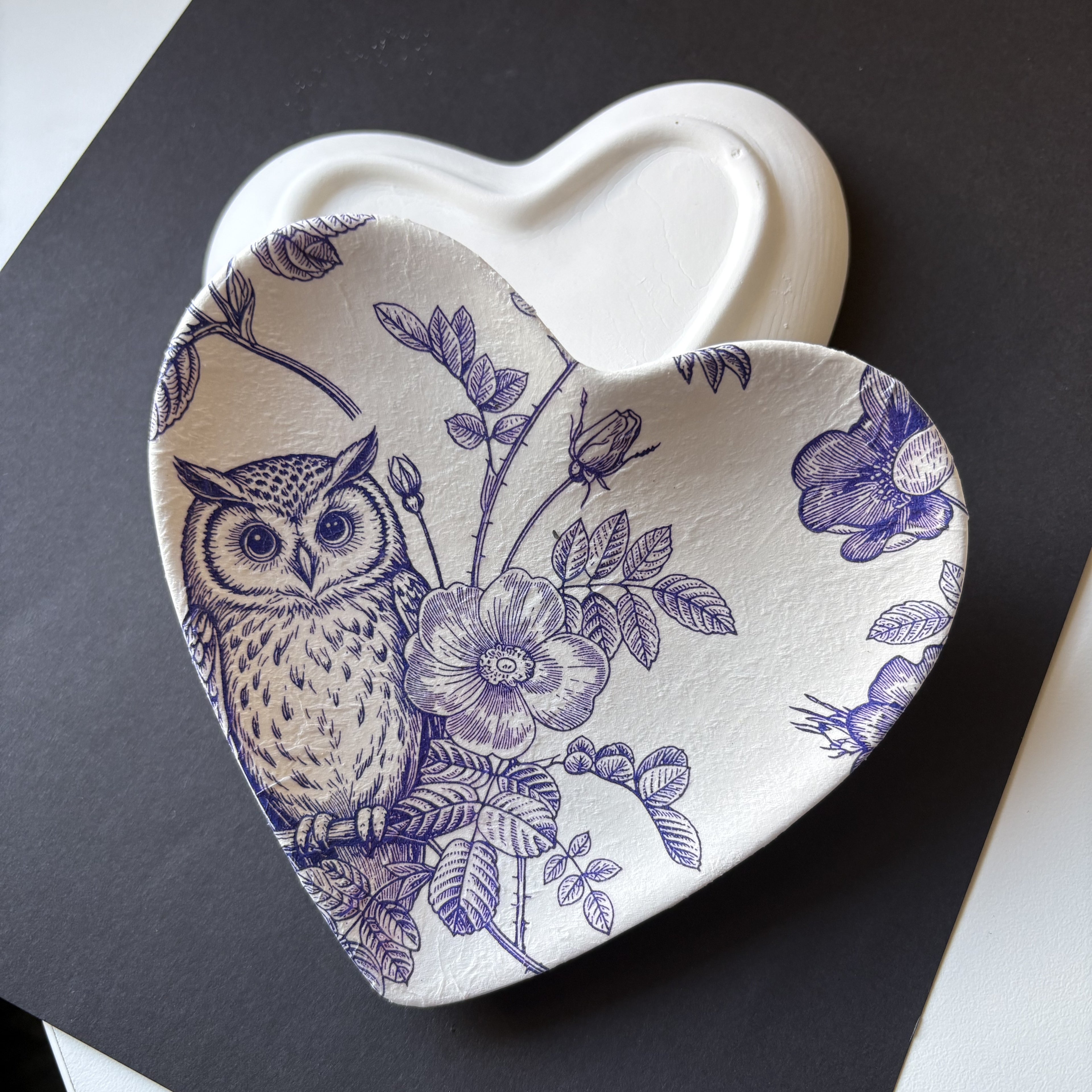 A heart-shaped ceramic dish with a blue floral and owl pattern, shown on top of a matching plain heart-shaped dish against a dark background.