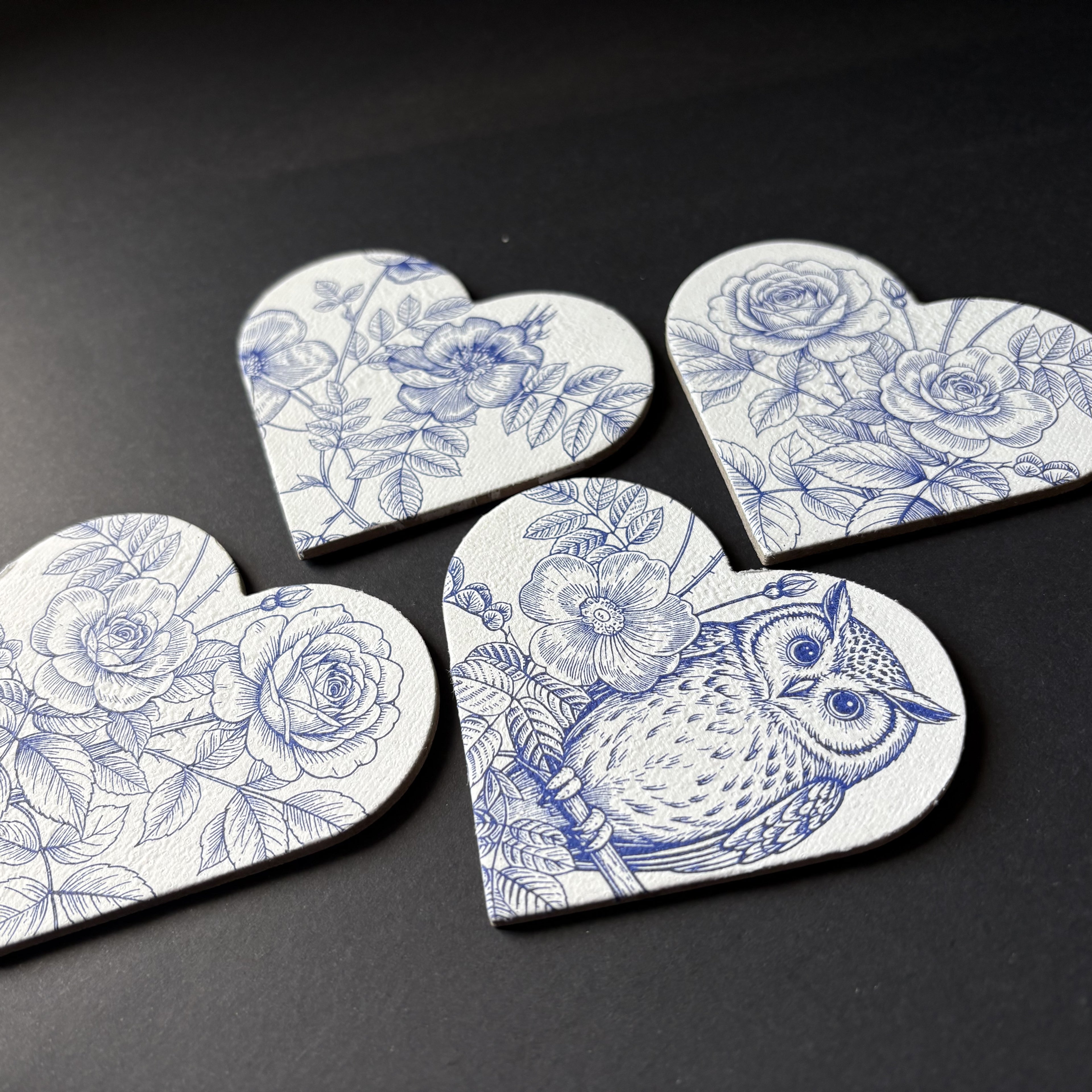 Four heart-shaped magnets with blue floral and owl illustrations on a white background, arranged on a dark surface.