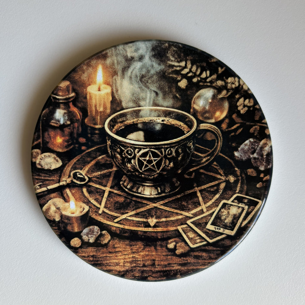 A round decorative coaster featuring a dark, mystical illustration of a steaming cup with a pentagram design, surrounded by candles, tarot cards, crystals, and a spellbook on a wooden surface, photographed against a plain light background.