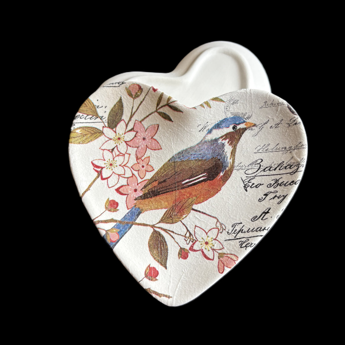 A heart-shaped ceramic dish with a vintage-style bird and floral print on a cream background, set against a solid black background.