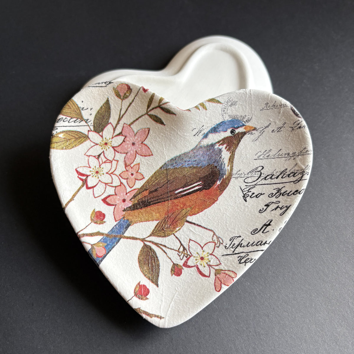 A heart-shaped ceramic dish featuring a vintage-style bird illustration with pink blossoms and cursive script on a dark gray background.