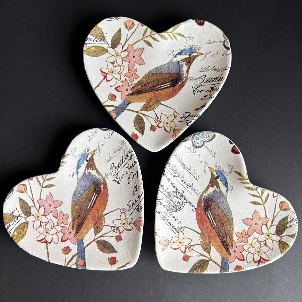 Three heart-shaped ceramic trinket trays arranged in a triangle on a dark background. Each tray features a vintage-style illustration of a bluebird perched on a flowering branch with script text in the background.