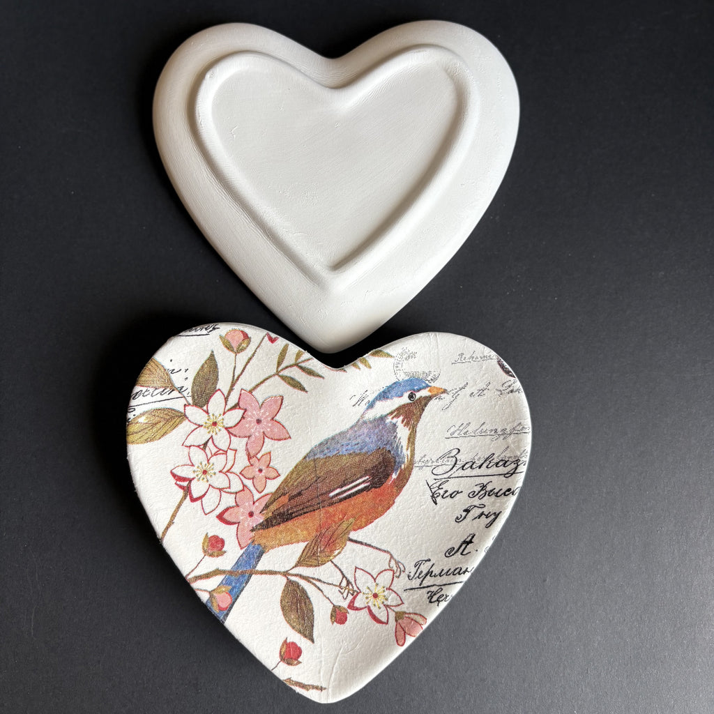 A heart-shaped ceramic dish with a white glazed top and a printed bottom featuring a bird, flowers, and script text, shown on a dark background.