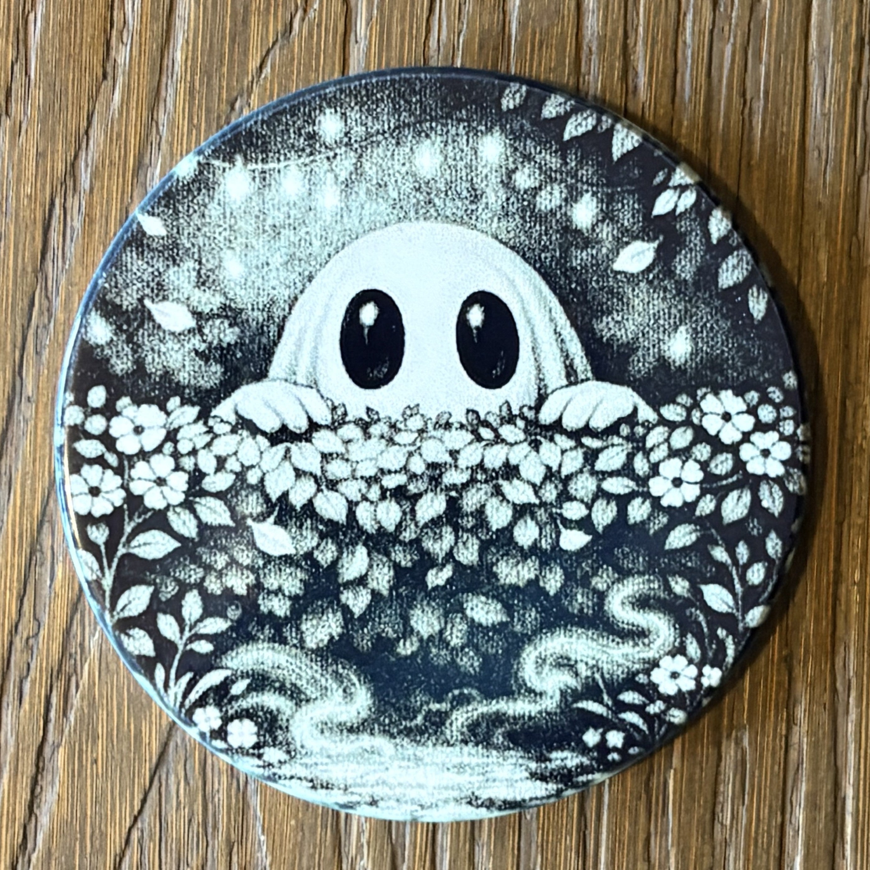 A round coaster featuring a black and white illustration of a cute ghost peeking over a bed of flowers, placed on a wooden surface.