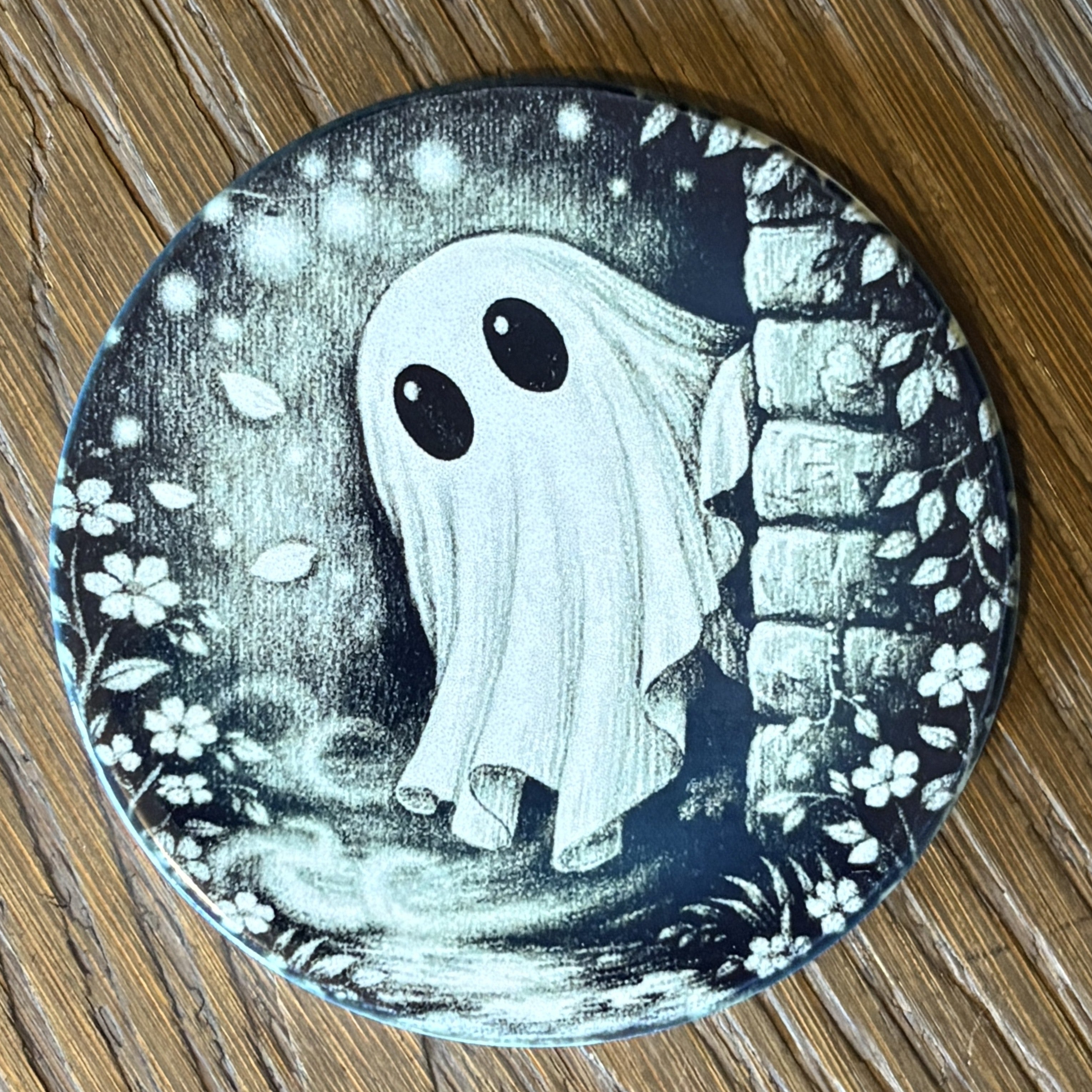 A round printed coaster or mat featuring a spooky cartoon ghost with large black eyes standing beside a stone wall, photographed on a wooden surface.