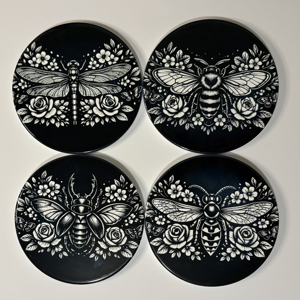 Four black coasters with white bee, dragonfly, wasp & beetle and rose designs on a light gray background.