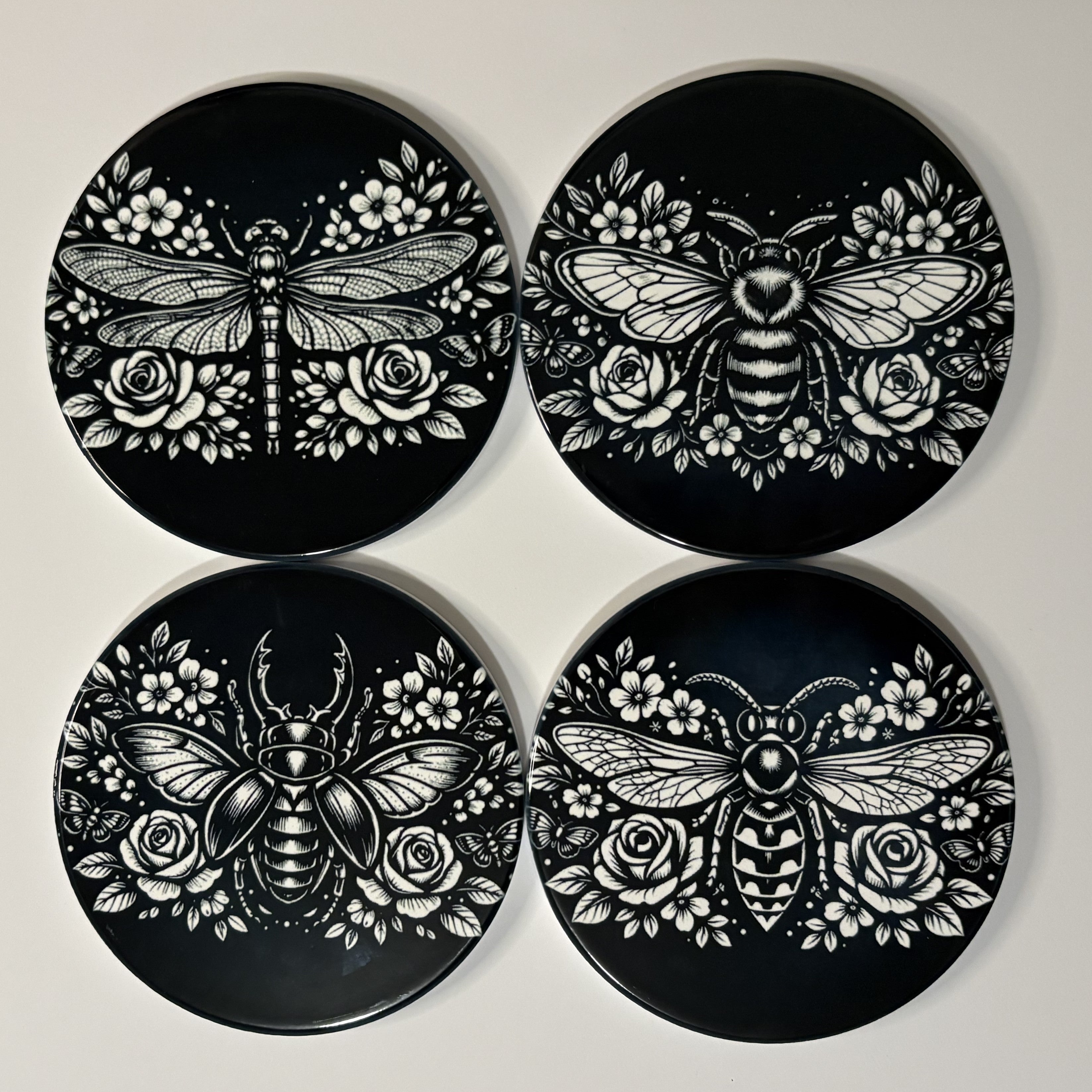 Four black coasters with white bee, dragonfly, wasp & beetle and rose designs on a light gray background.