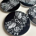 Four black coasters with white bee, dragonfly, wasp & beetle and rose designs on a light gray background.