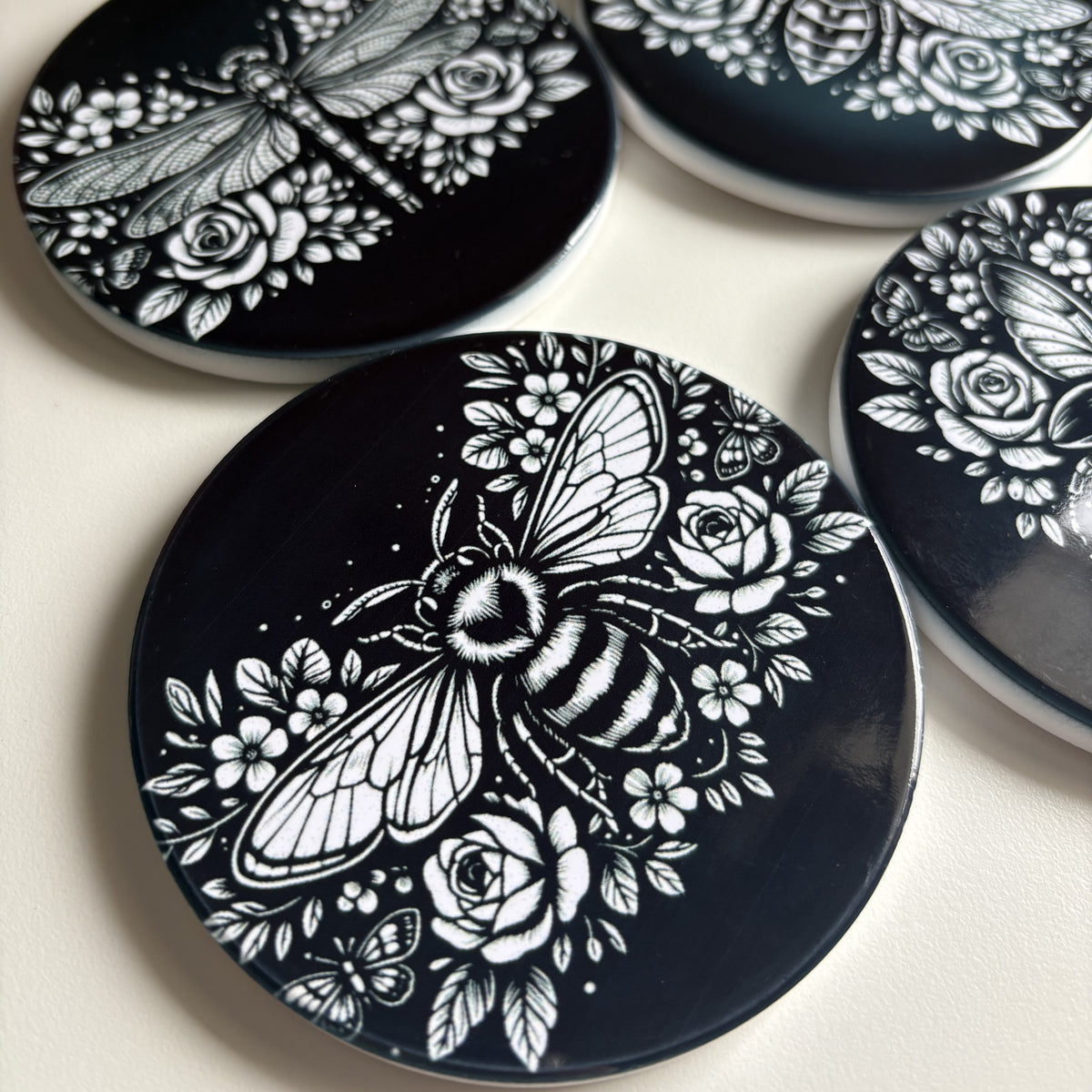 Four black coasters with white bee, dragonfly, wasp & beetle and rose designs on a light gray background.