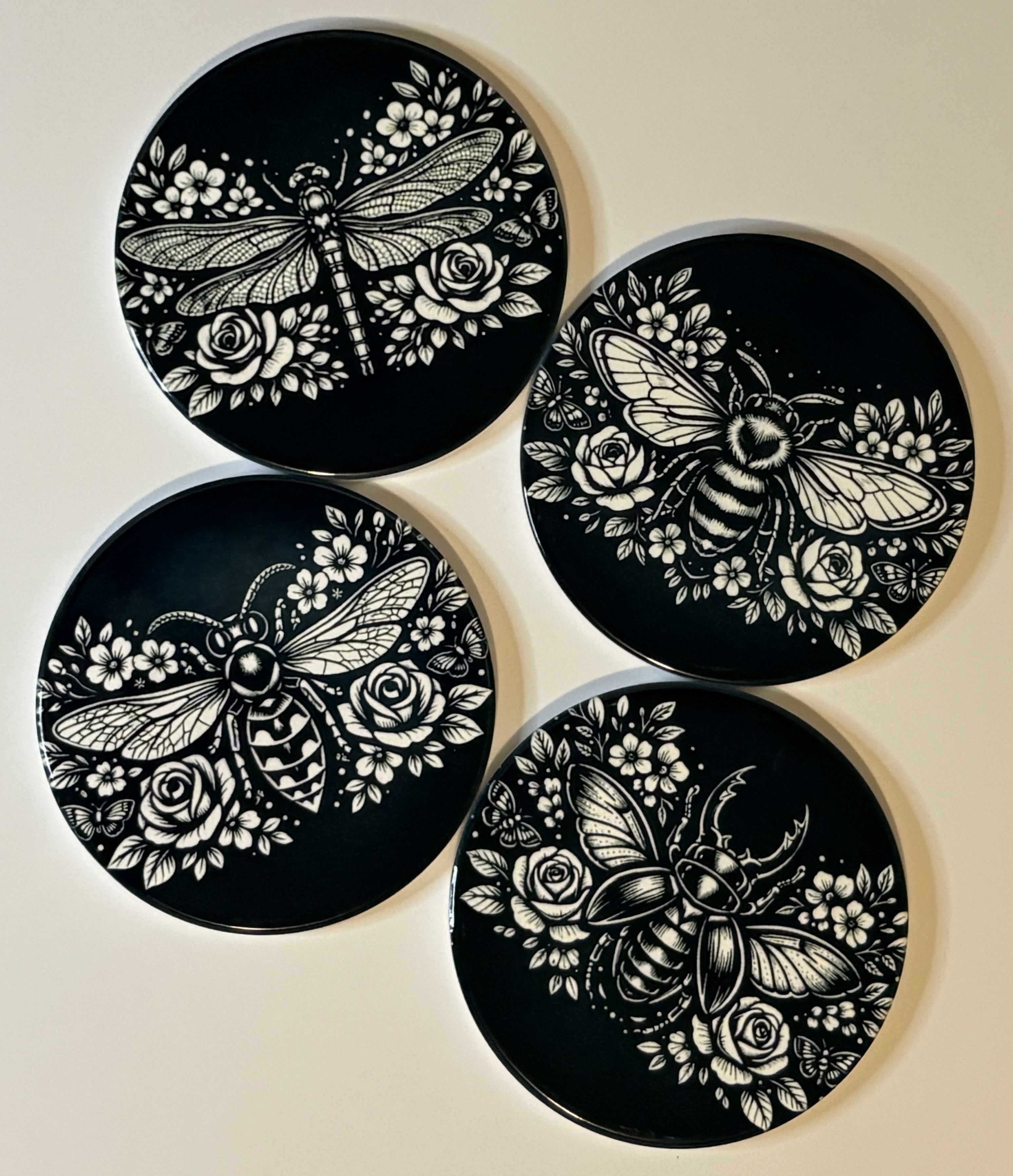 Four black coasters with white butterfly and floral designs on a beige background