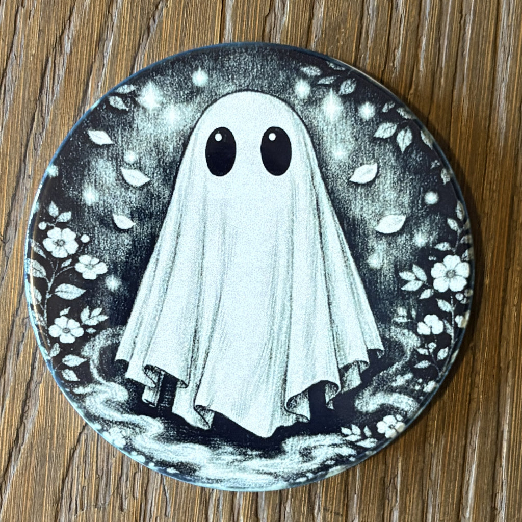 A round printed coaster or tile featuring a black and white illustration of a cute ghost with large eyes, surrounded by leaves and flowers, placed on a wooden surface.