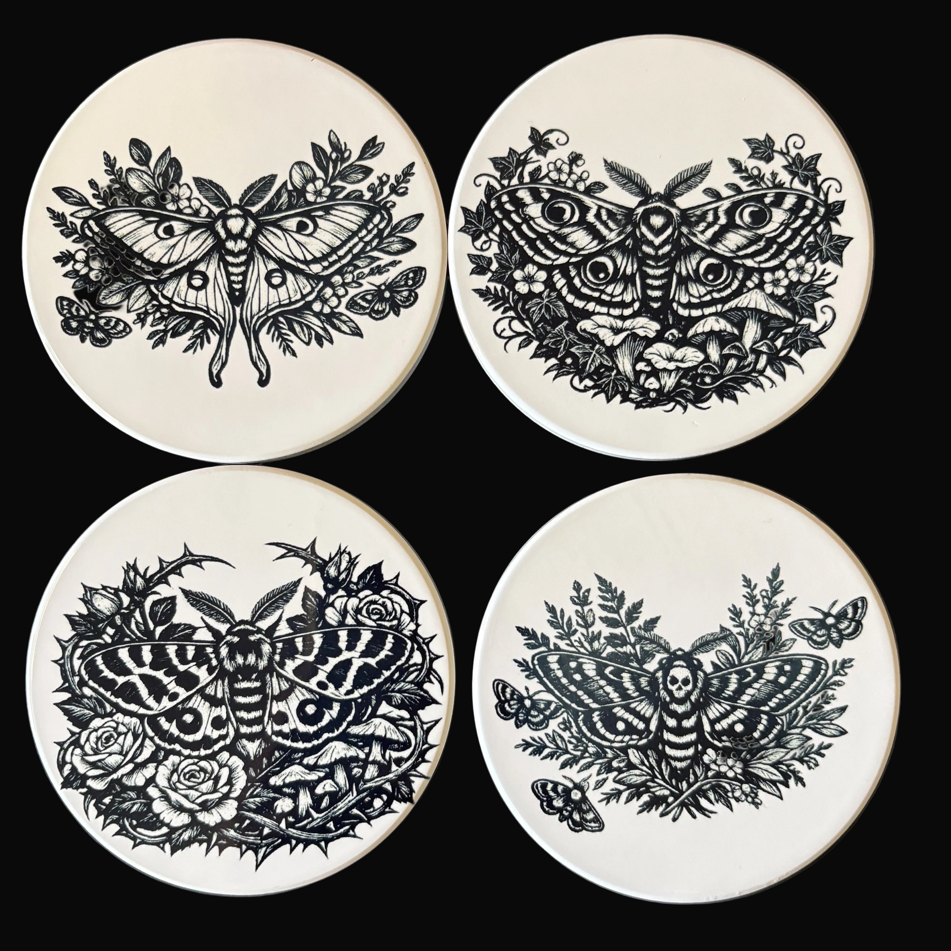 Four ceramic coasters with black moth designs on a dark background
