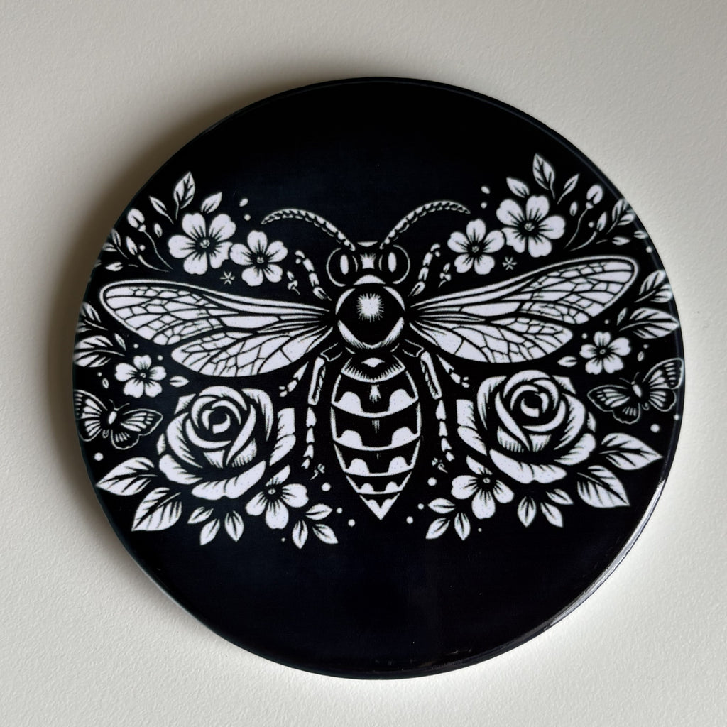 A round black coaster featuring a detailed white graphic of a bee surrounded by flowers and leaves, photographed against a plain light background.
