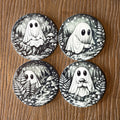 Four round coasters featuring black and white illustrations of cute ghosts in a forest, arranged in a 2x2 grid on a wooden surface.