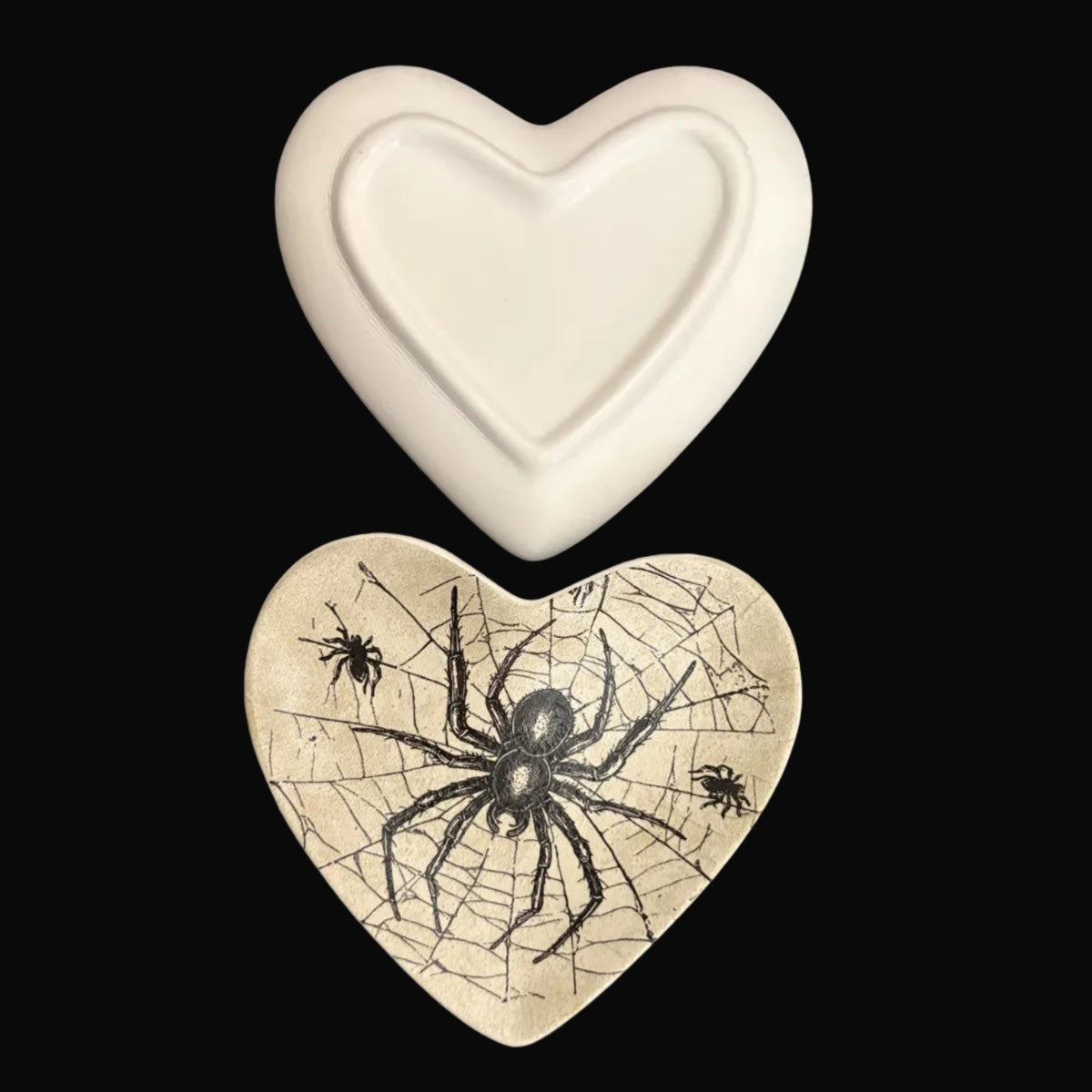 Two heart-shaped ceramic dishes or plates displayed on a dark background. The top plate is solid off-white, while the bottom plate is beige with a black spider and cobweb design.