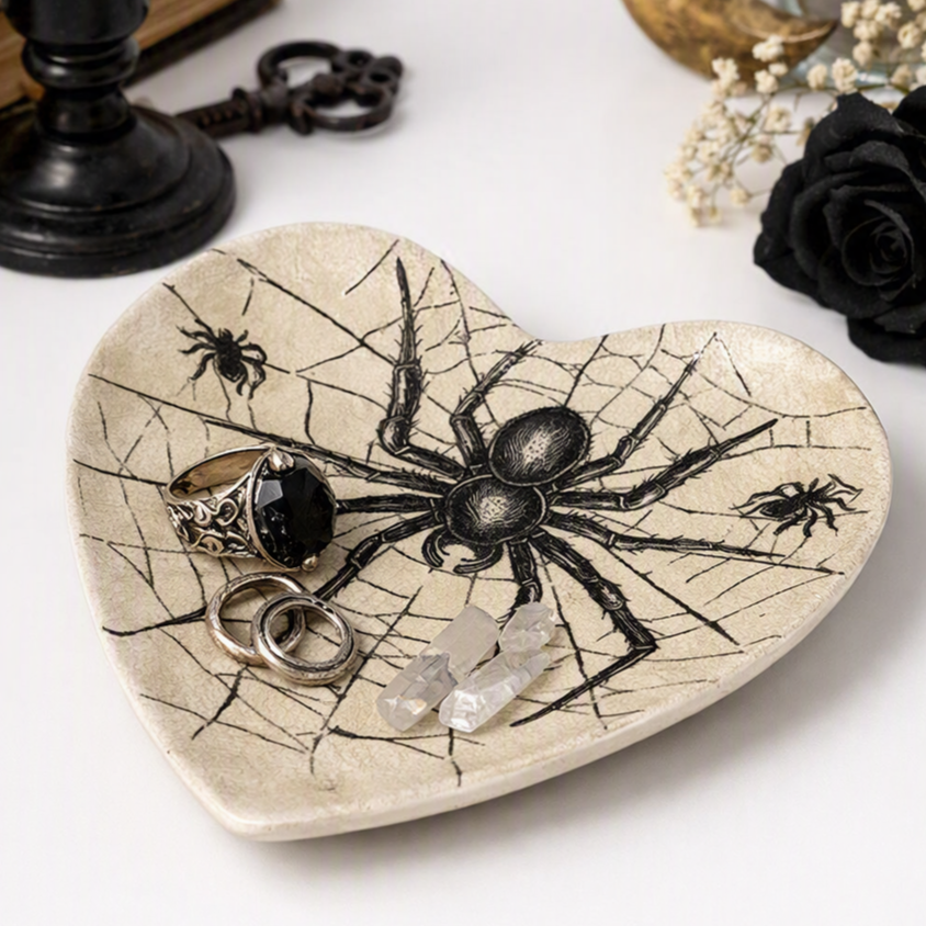 A heart-shaped ceramic jewelry organizer tray with a black spider and web design, styled with rings and crystals on a white surface with gothic props.