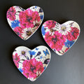 Three heart-shaped ceramic trinket tray with a vibrant floral print featuring pink, red, blue, and white flowers, arranged on a dark gray background.