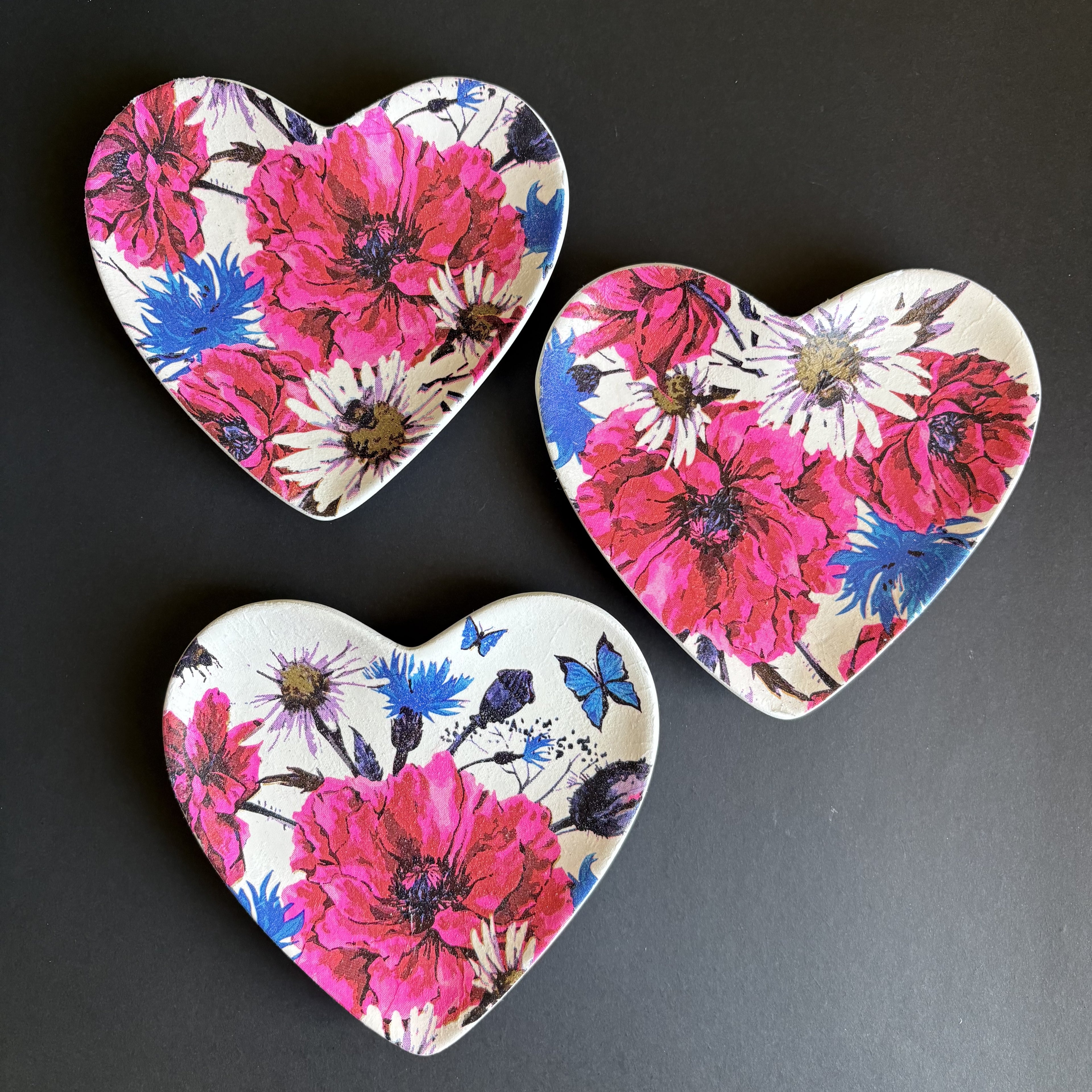 Three heart-shaped ceramic trinket tray with a vibrant floral print featuring pink, red, blue, and white flowers, arranged on a dark gray background.