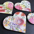 Heart-shaped magnet featuring a vibrant, multicolored floral print with pink protea-like flowers and green leaves on a white textured background, arranged on a dark surface.