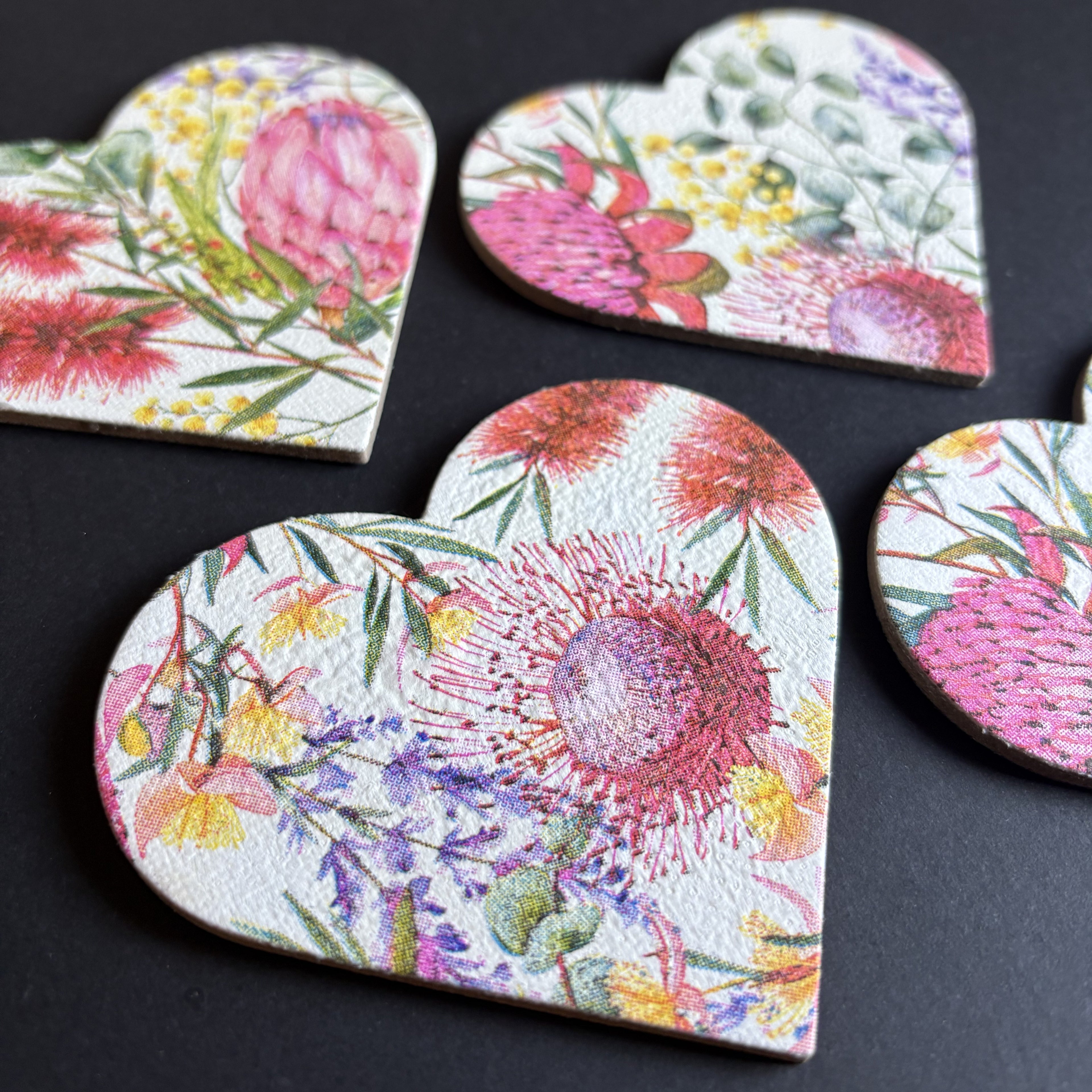 Heart-shaped magnet featuring a vibrant, multicolored floral print with pink protea-like flowers and green leaves on a white textured background, arranged on a dark surface.