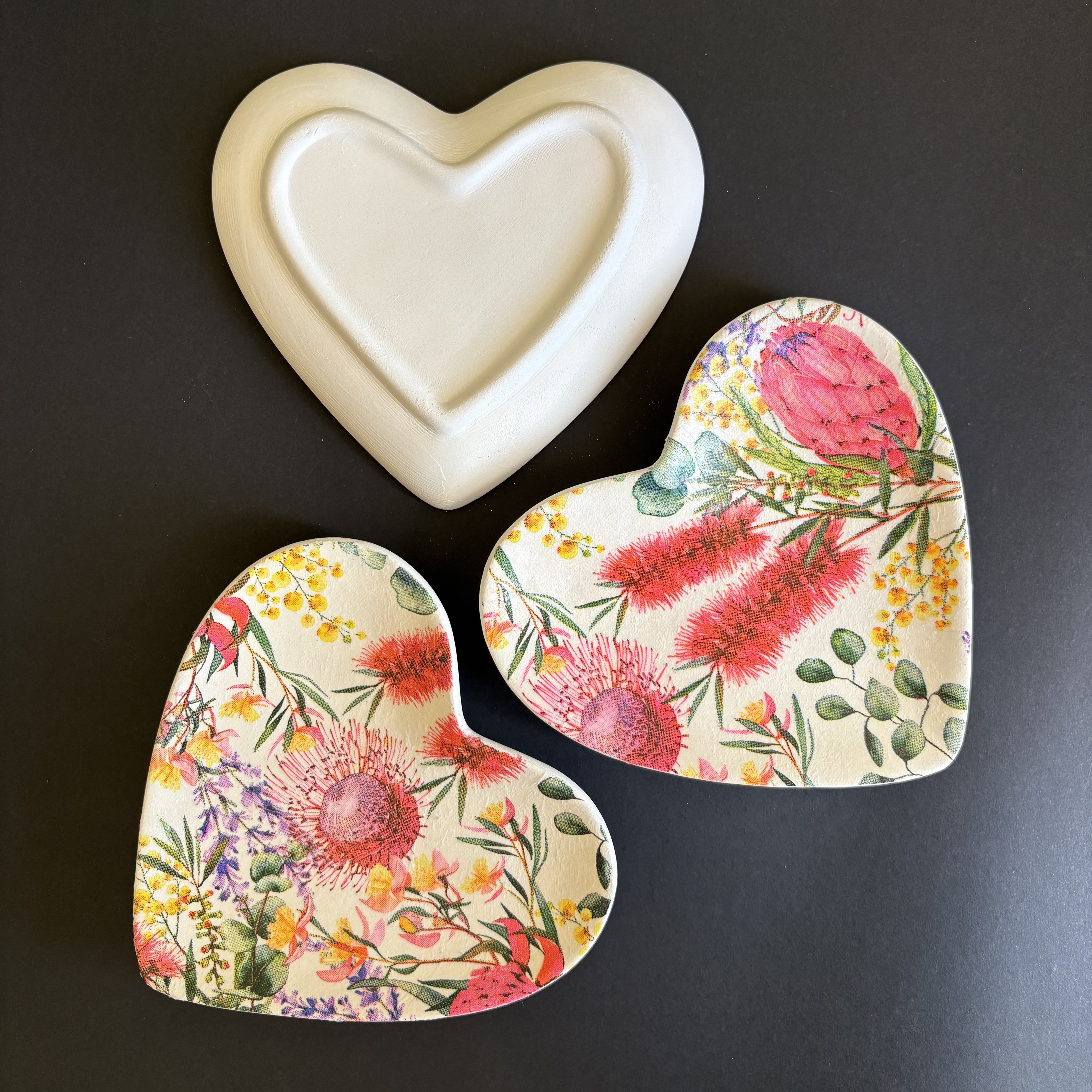 Three heart-shaped ceramic trinket tray with floral designs are arranged on a dark gray background. One trinket tray is solid cream-colored, while the other two feature a detailed botanical pattern with pink, red, yellow, and green flowers on a cream base.