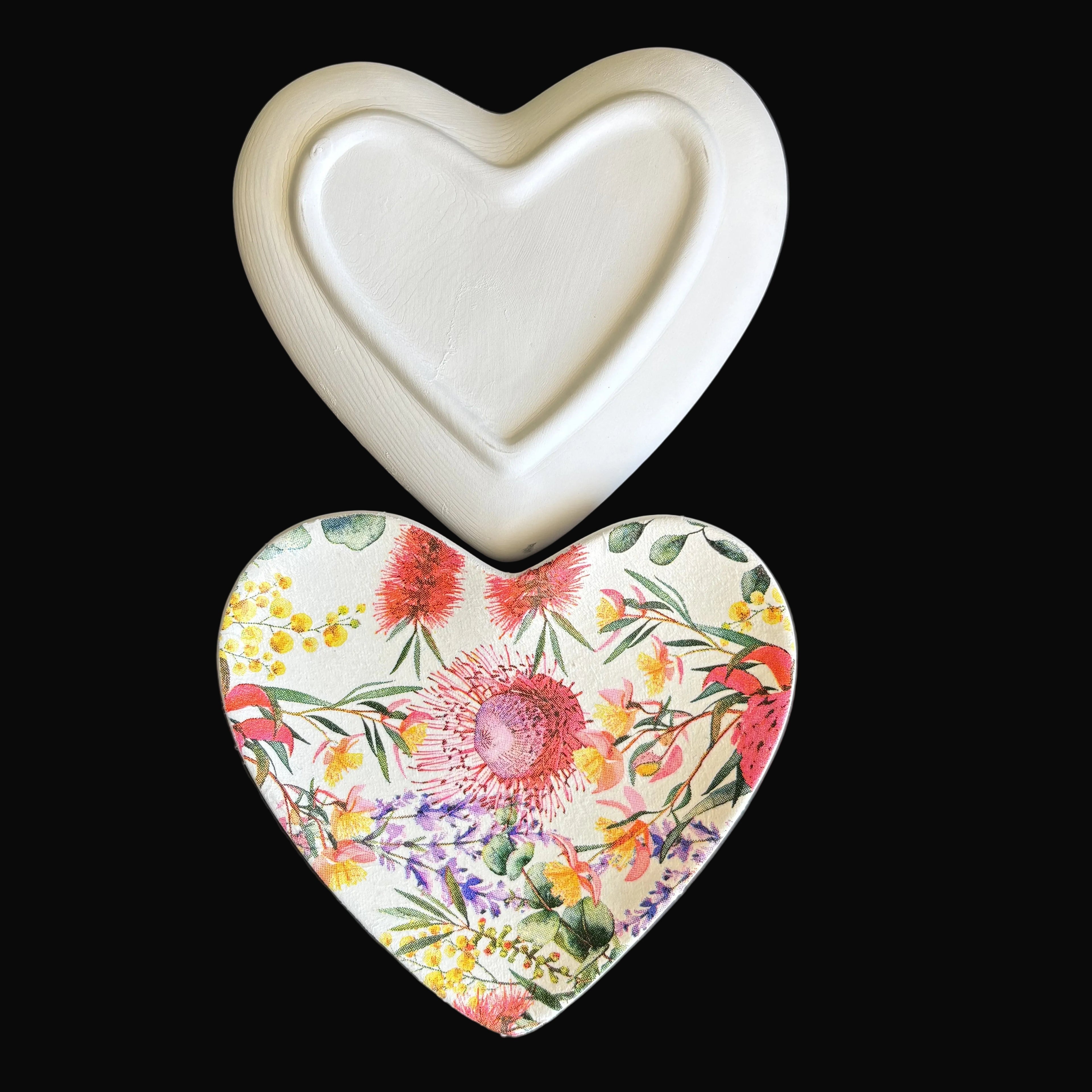 Two heart-shaped ceramic trinket tray on a dark gray background. The top dish is solid matte white, and the bottom dish features a colorful floral print with pink, yellow, and red flowers on a cream background.