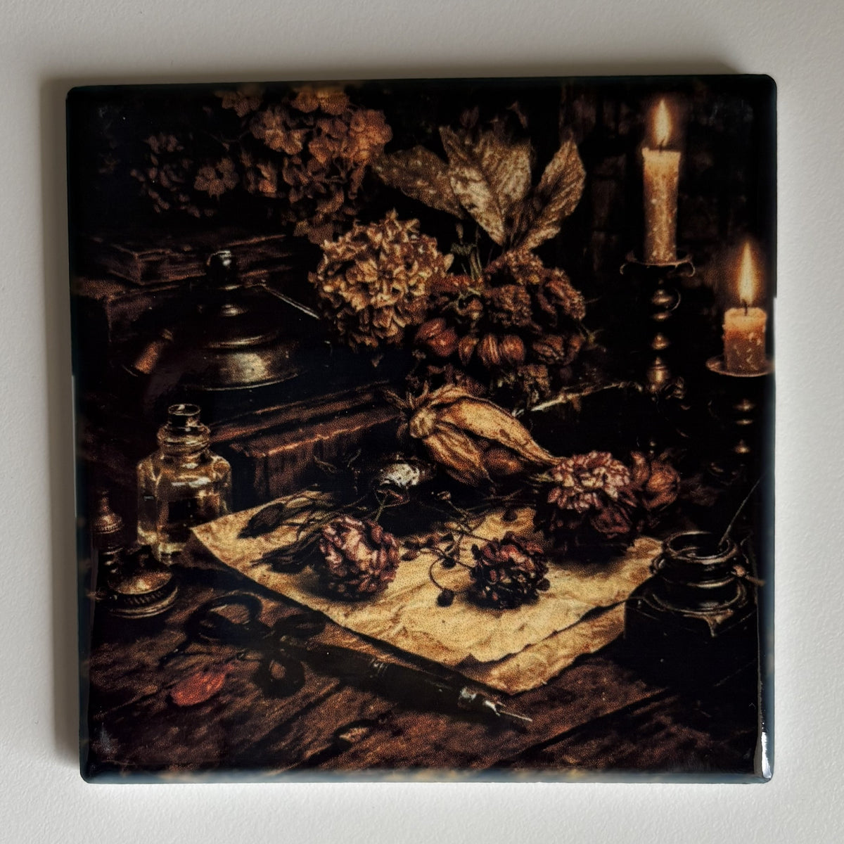 A square coaster featuring a dark, moody still life print with dried flowers, candles, and vintage objects on a wooden table, shown against a plain light gray background.