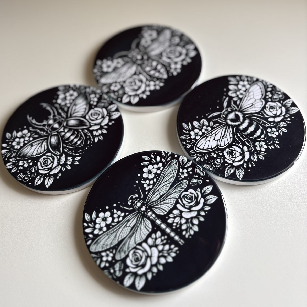 Four round black coasters with white floral and insect illustrations, arranged on a light beige surface.