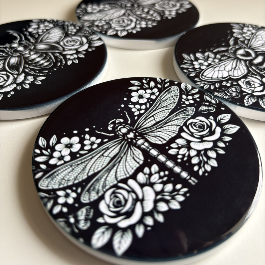 A close-up of round black coasters with white floral and insect illustrations, including a dragonfly, bee, and butterfly, arranged on a light surface.