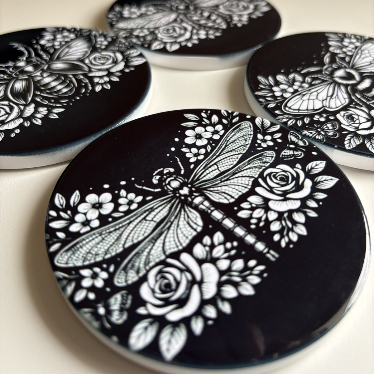 A close-up of round black coasters with white floral and insect illustrations, including a dragonfly, bee, and butterfly, arranged on a light surface.