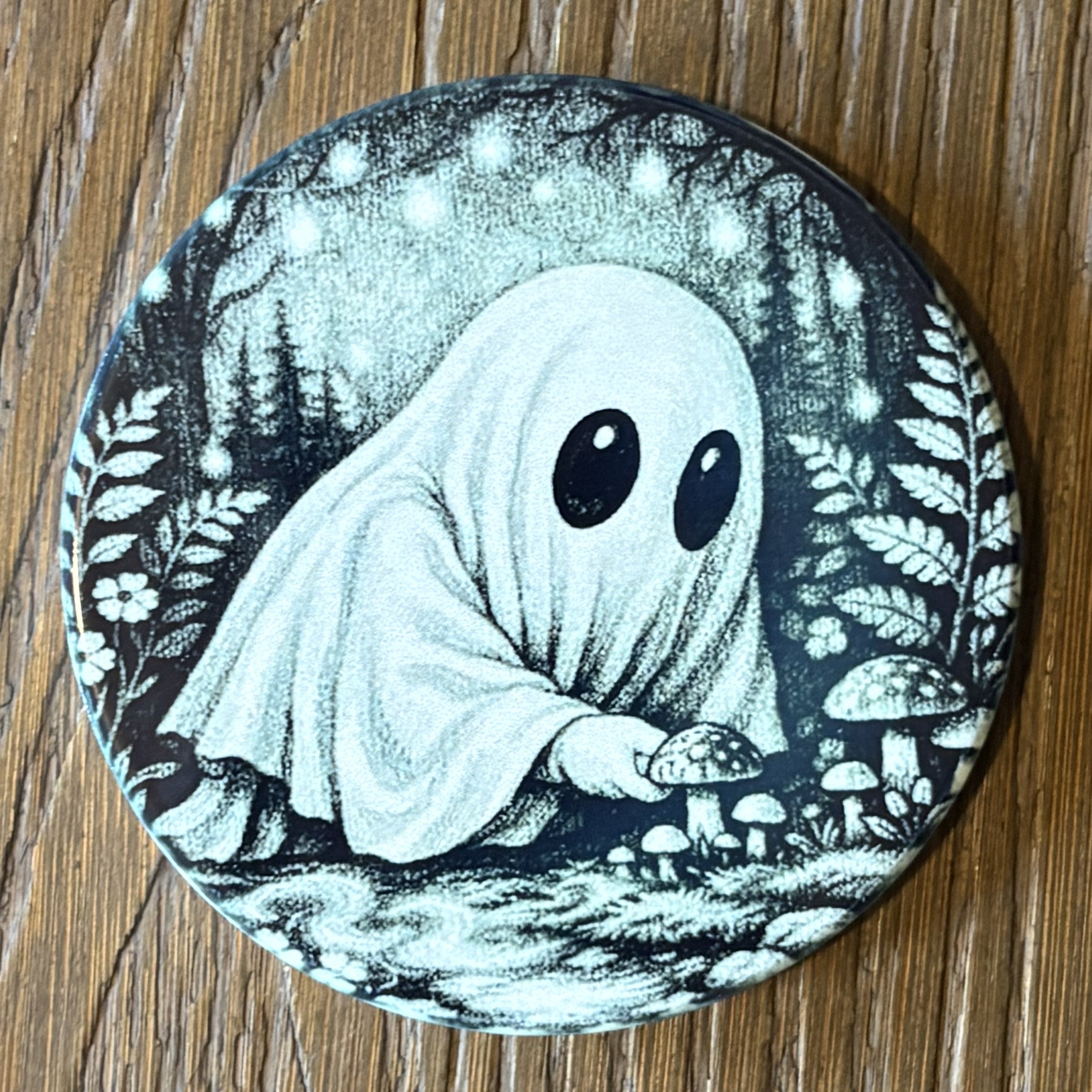 A round coaster featuring a black and white illustration of a cute ghost with large eyes sitting among mushrooms in a forest, placed on a wooden surface.