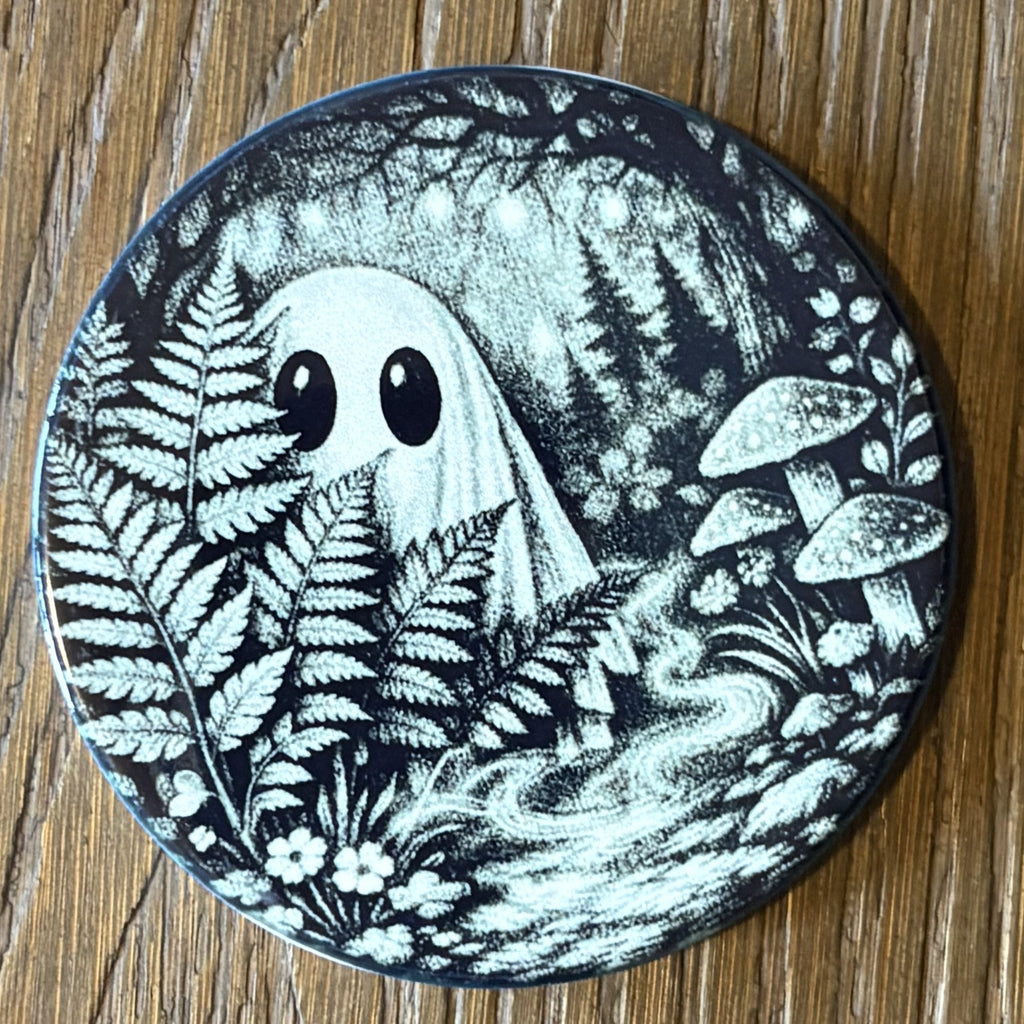 A round coaster featuring a black and white illustration of a cute ghost in a forest with ferns and mushrooms, placed on a wooden surface.