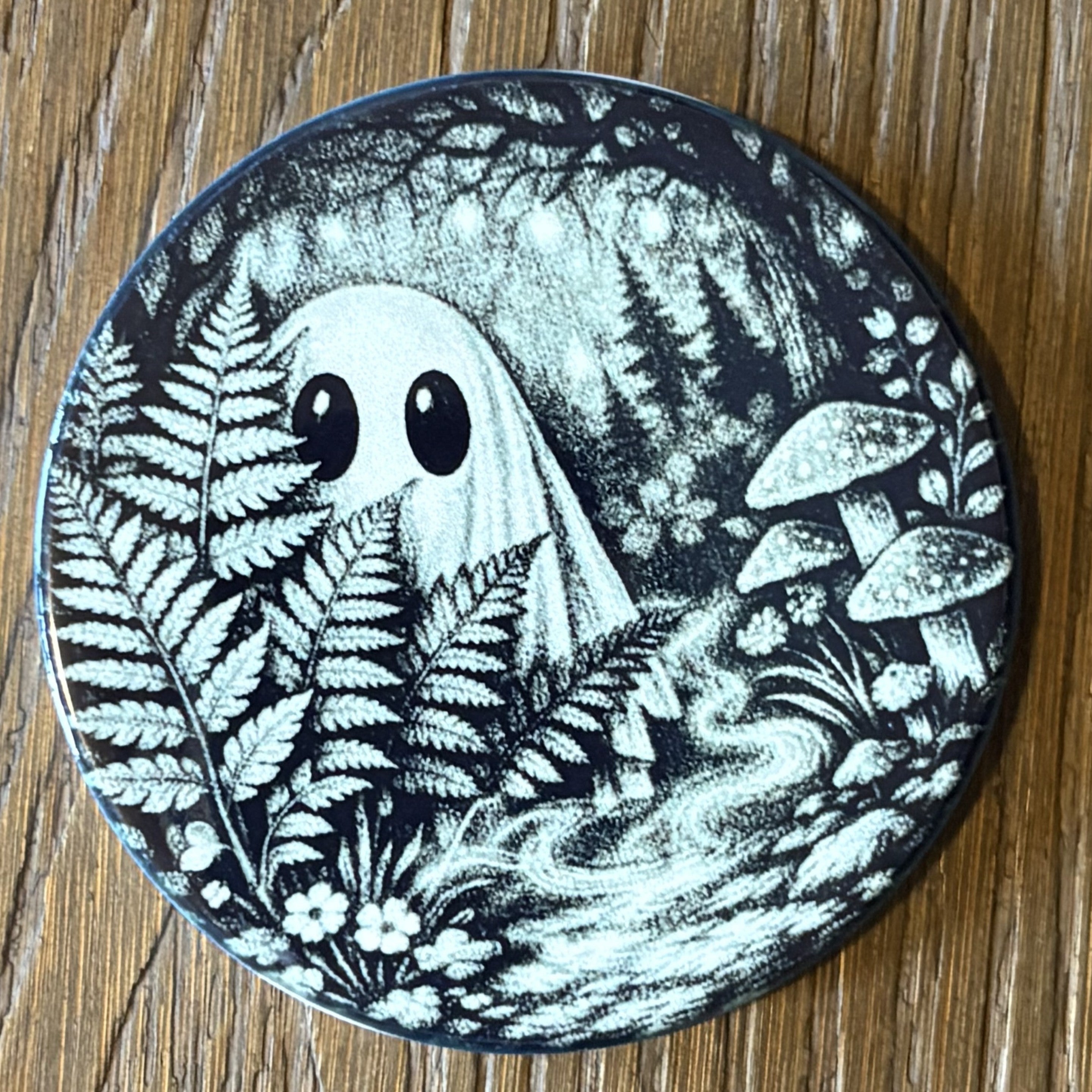 A round coaster featuring a black and white illustration of a cute ghost in a forest with ferns and mushrooms, placed on a wooden surface.