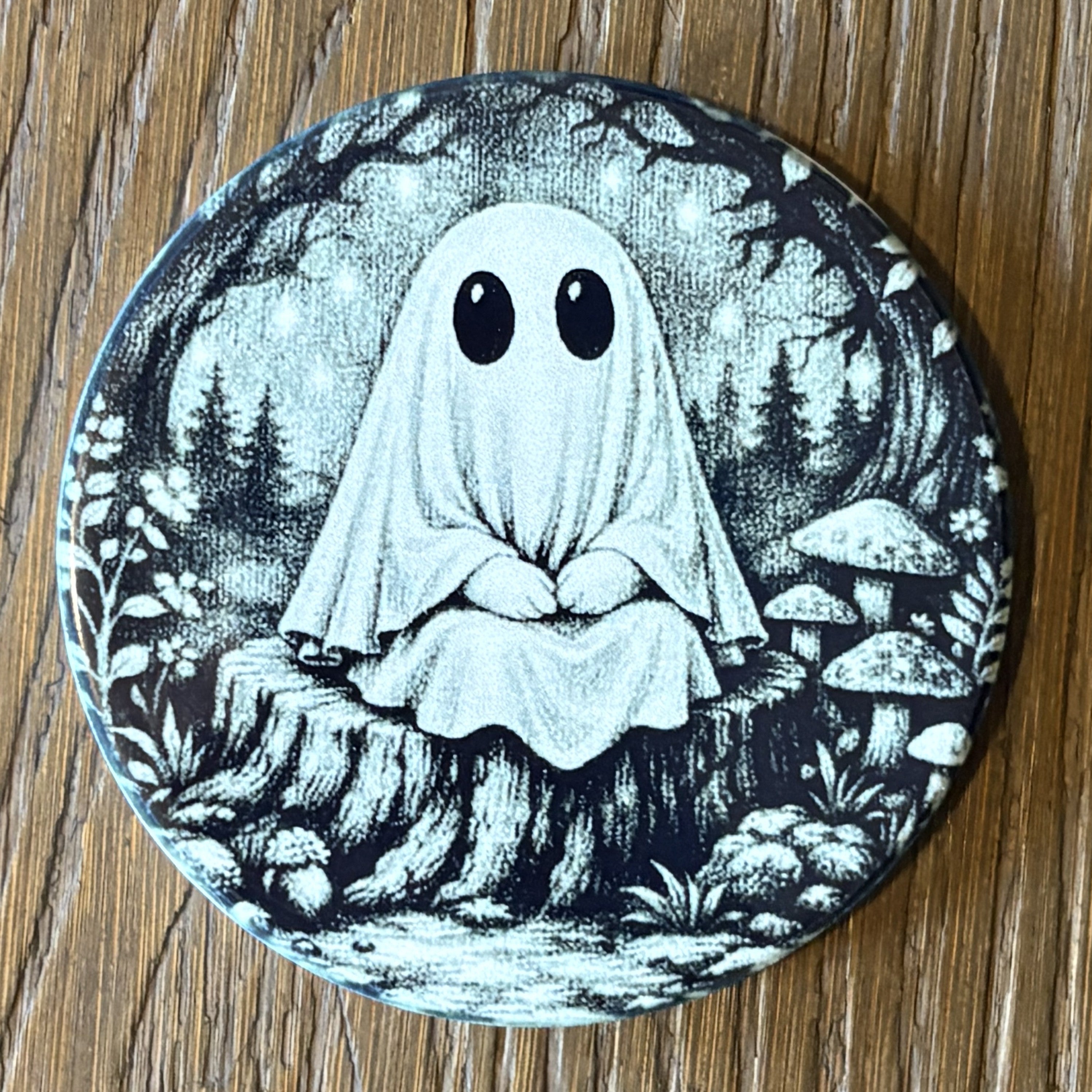 A round coaster featuring a black and white illustration of a ghostly figure sitting cross-legged in a forest with mushrooms, placed on a wooden surface.